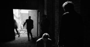 A man stands menacingly in front of a defeated, restrained man in a dark garage.