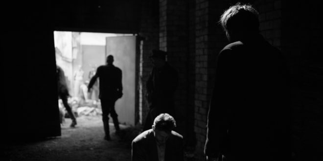 A man stands menacingly in front of a defeated, restrained man in a dark garage.