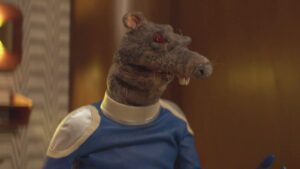 A rat puppet with red eyes wearing a blue and yellow shouldered shirt