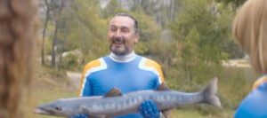A wet man wearing a blue and yellow Super Sentai suit proudly holds a 2 foot long barracuda.